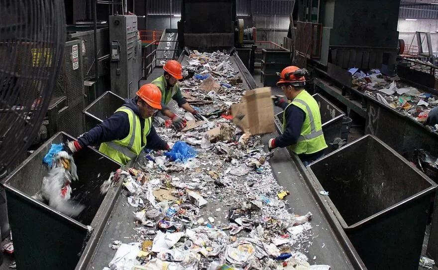 Workers sorting mixed recyclables by hand on a conveyor belt inside a recycling facility