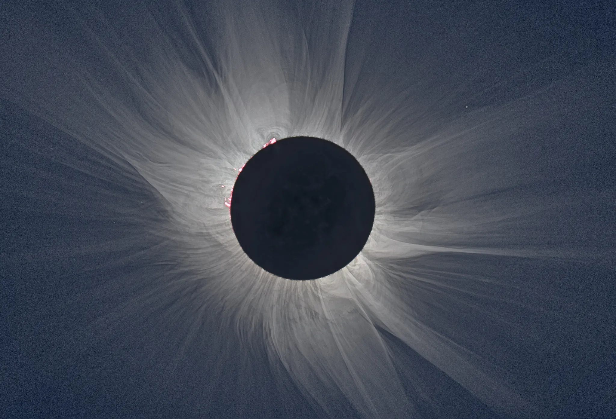 White solar corona streaming around the dark disk of the Moon during totality, with faint red prominences near the edge.