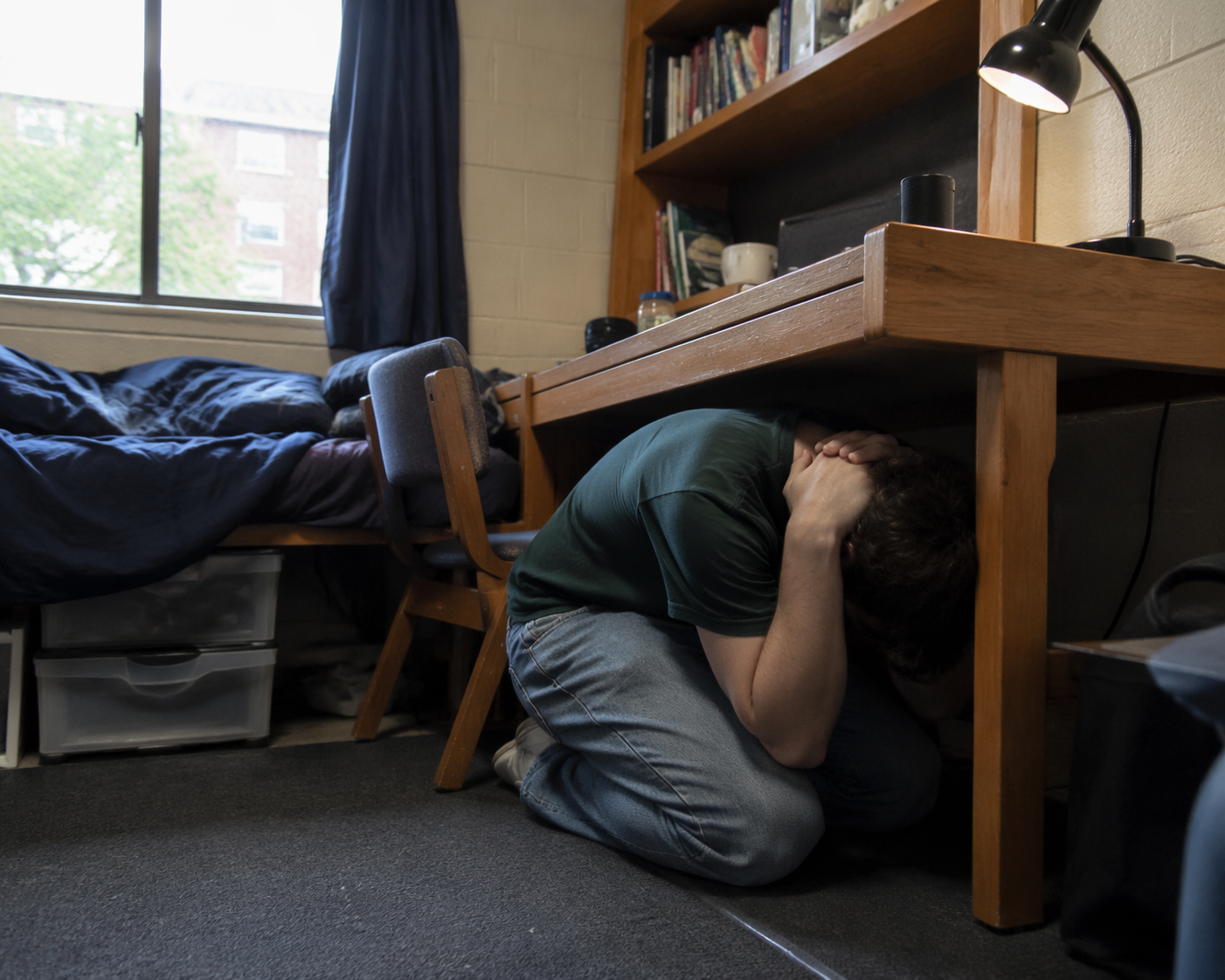 A college student crouched under a sturdy desk in a cramped dorm room in the Drop, Cover, Hold On position, with a bed, bookshelf, and window nearby