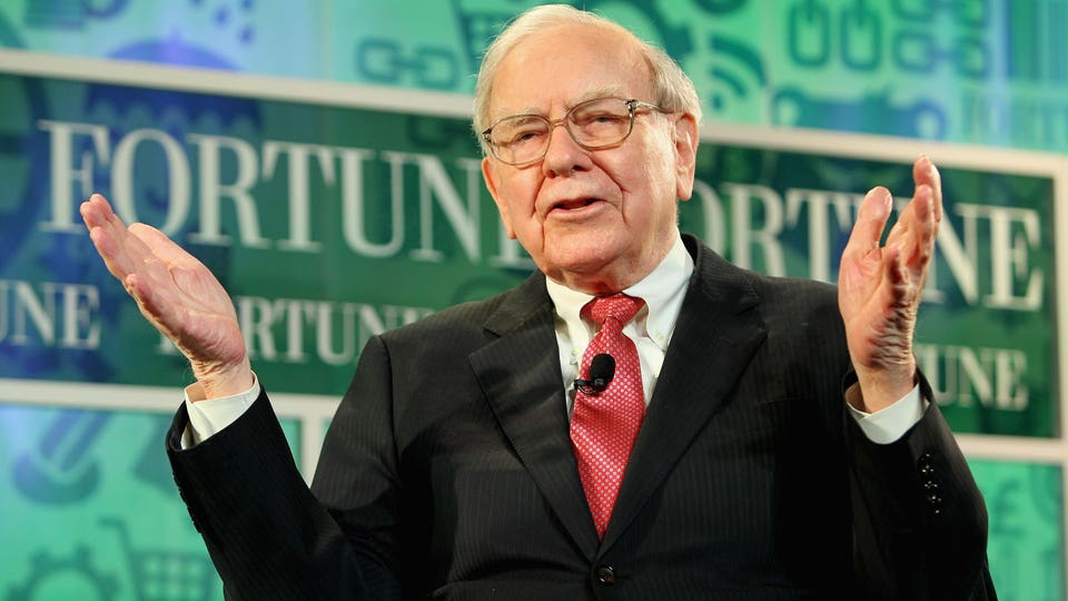 Warren Buffett speaking at a podium in a dark suit and glasses