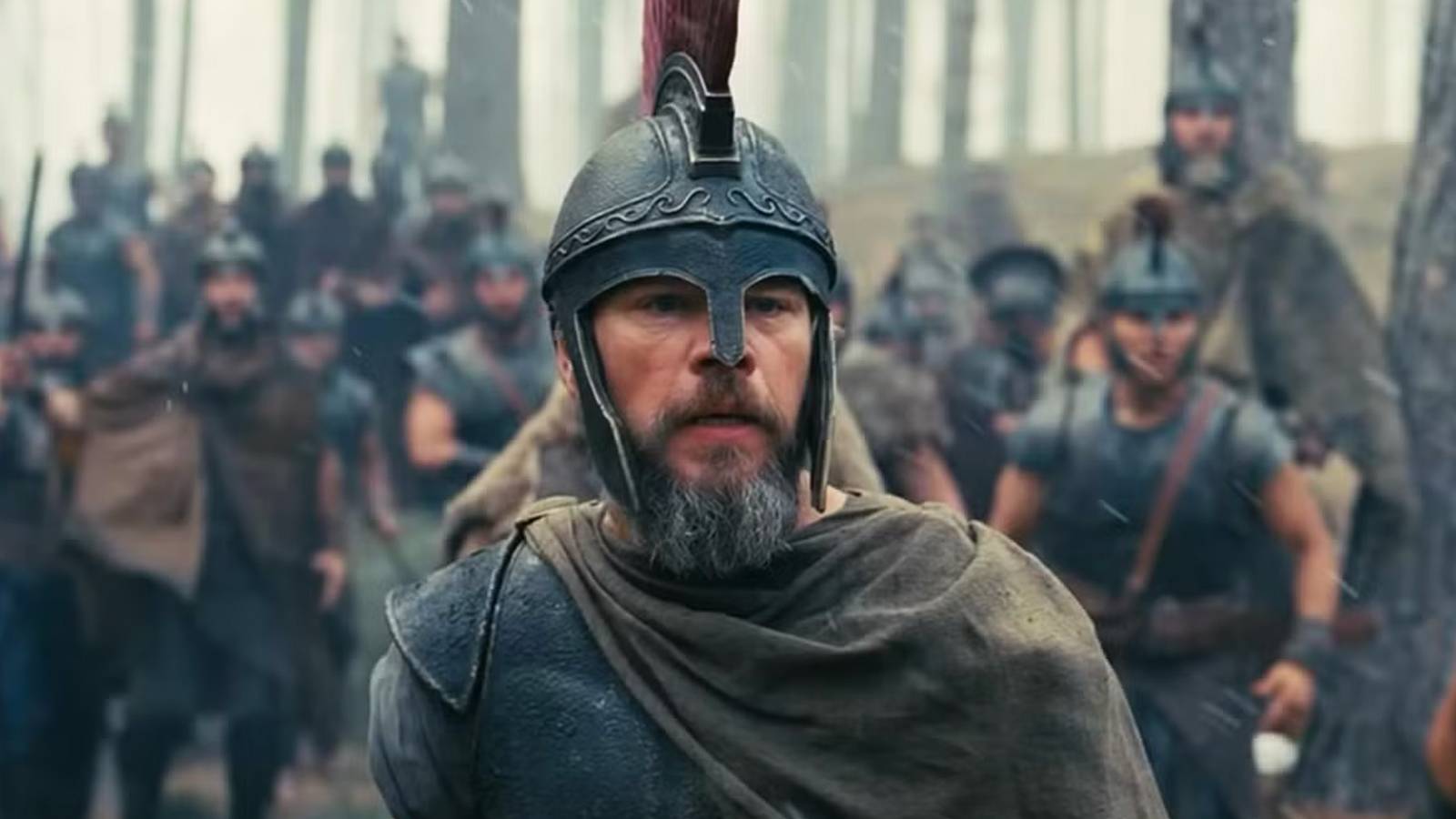 Matt Damon as Odysseus standing in armor before armed soldiers at dusk